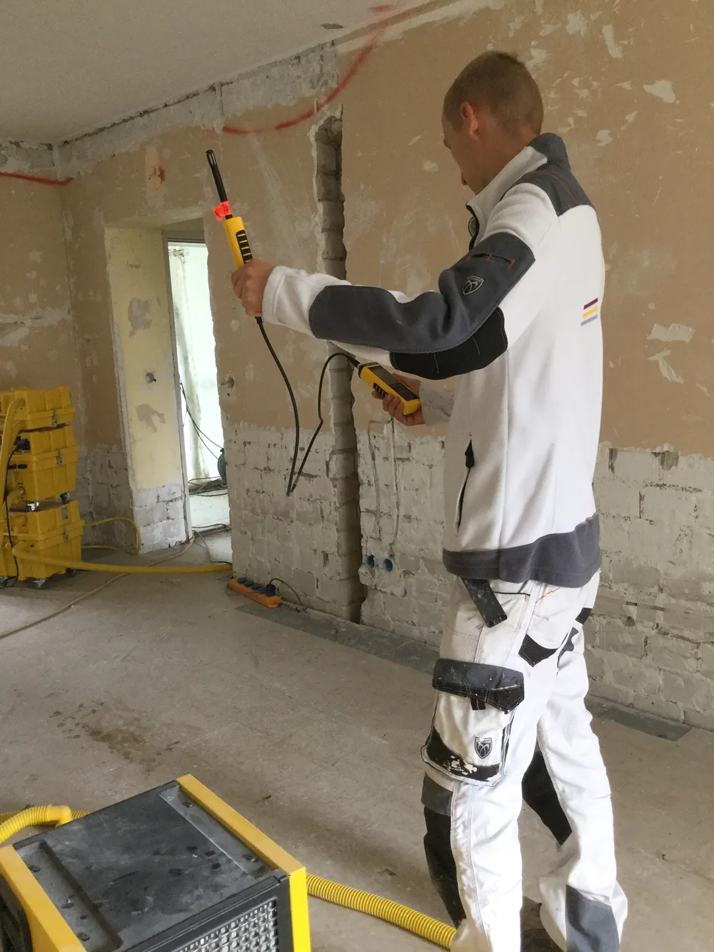Feuchtemessung an einer zurückgebauten Wand mit Messgerät, im Hintergrund ein Bautrockner.